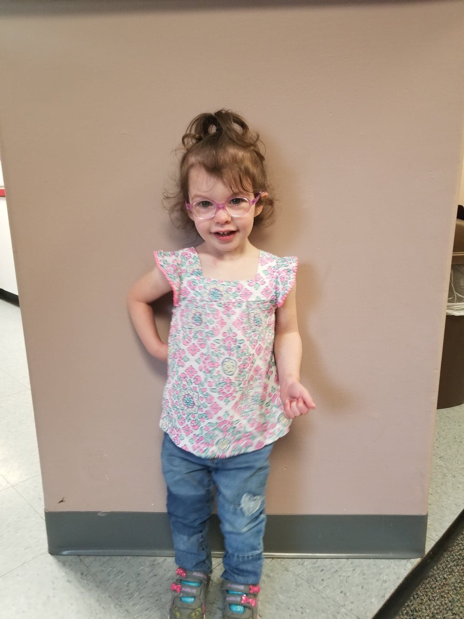 A young girl with glasses stands in front of a wall
